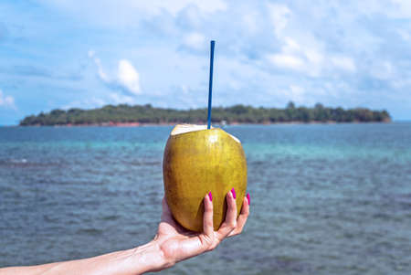 Fresh coconut cocktail in hand on turquoise sea background, travel card. Women's hand with pink nail Polish holding a coconut with a tube on the background of the island in the blue sea.の写真素材