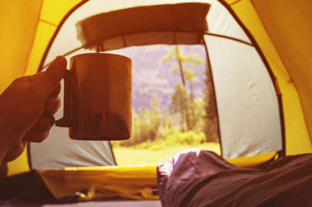 traveling and spending time in tent while drinking coffee in the morning. View from inside. Camper holding mug with hot tea inside the tent in the background of a beautiful mountain landscape.の写真素材