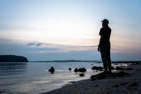 Silhouette of alone man looking toward vibrant sunset. Flare and reflected in shallow waters of sea levelの写真素材