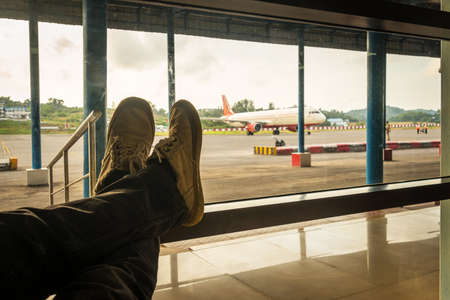 male waiting for her flight. The passenger sits at the airport with his leg folded against the background of aircraft on the runway.の写真素材