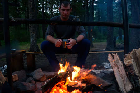 a man looks at the fire. Young man sitting on the bench and drinking tea while looking at fire. Tourist Hiking romance.の写真素材