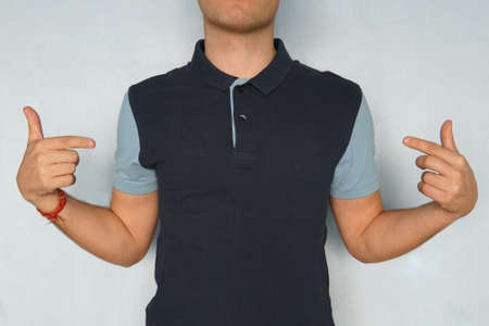 Design and advertising concept. Cropped shot of stylish young man pointing index finger at copy space on his casual t-shirt, standing indoors against blue wall, point a finger,の写真素材