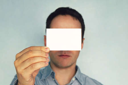 young man holding visit card. businessman closes his eyes business card. Blank for text. White business card in hand. Horizontal Mockupの写真素材