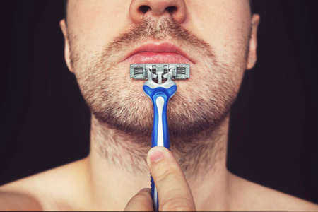 Young man without shaving cream on his face, grooming his beard with straight razor on black backgroundの写真素材