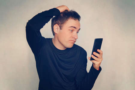 A thoughtful young man reads a message on a black modern smartphone. Indifferent emotion on his face. Guy scratching his head looking at the screenの写真素材