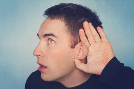 Close up portrait of curious interested delightful funny amazed cheerful surprised man holding hand near ear and trying to hear the information on blue backgroundの写真素材