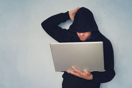 thoughtful Hacker with his computer thinking on blue background.. hacker in a black hood with a gray...