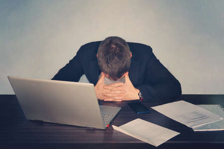 Exhausted tired businessman working on laptop at office, massaging temporal area, holding glasses, feeling fatigue discomfortの写真素材