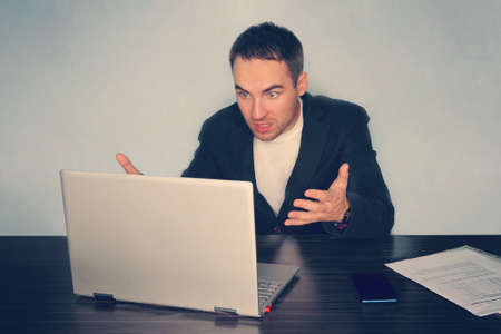 Frustrated young businessman looks at laptop screen screaming in anger at workplace in office. Unexpected high bill, unpaid debt, failing financial report tax delinquency, breach of contract concept.の写真素材