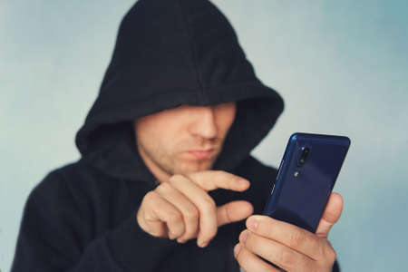 Faceless unrecognizable hooded person using mobile phone, identity theft and technology crime concept, selective focus on body. hacking a smartphone. identity theft.の写真素材