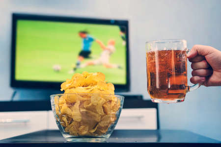Beer mug with beer in hand a man next to the chips on the background of a large TV on the screen which is a football matchの写真素材