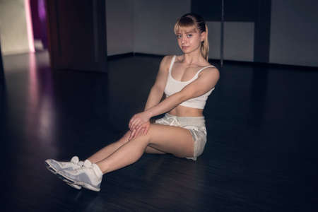 Happy cute young sportswoman sitting over dark background. Blonde athlete with shorts and a white...