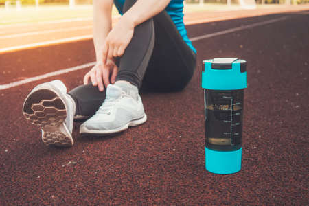 Young girl sitting down with protein shake or water bottle on street after running exercise running workout street outdoor in summer for muscle building. rest after morning runの写真素材
