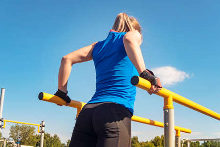 Fit young woman holding balance on the parallel bars. Blonde girl trains outdoors in the Park. Training of biceps and triceps.の写真素材