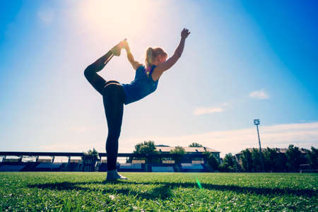 Young sportswoman doing warm-up balance exercise standing on one legs stretching leg and back outdoors. Warm-up stretching at the stadium.の写真素材