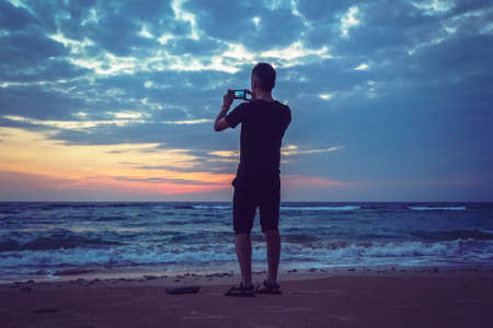 Silhouette of man keep memories with camera phone in his hand. Marvelous sunset at sea, smooth water levelの写真素材