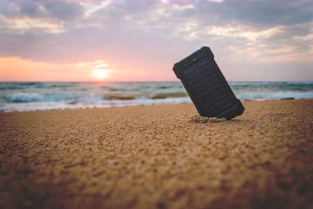 Powerbank in the sand on the background of the rising sun in the cloudy sky and the blue sea. Alternative energy source. battery is charged by solar energy. on desert island. beachの写真素材