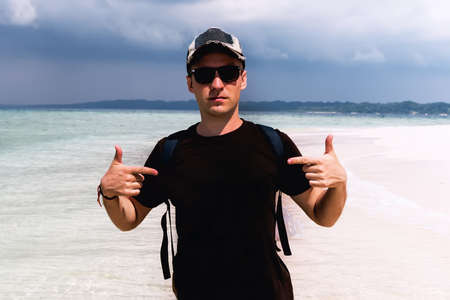 A tourist in a cap and sunglasses. Happy man indicating himself against the background of the sea landscape on an exotic island. copyspace, copy spaceの写真素材