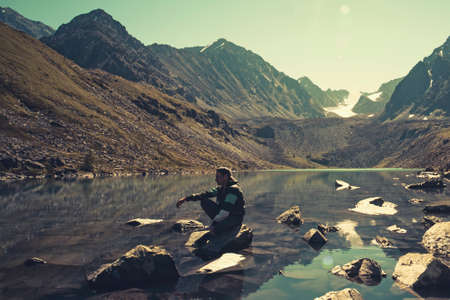 Calm man sitting on a rock on vacation looking at the lake and mountains on a summer evening. Lake...