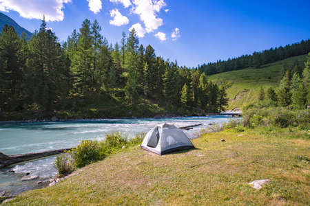 white tent on the grassy shore of a mountain lake amid snow-capped peaks under the clouds and blue sky lake, Altai mountains, Siberia, Russia. summer holidays in nature. wildlifeの写真素材