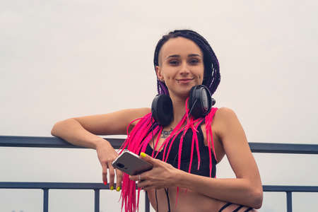 Beautiful young woman with mobile phone outdoors. Woman with piercing and African pink pigtails listening to music in black headphones. subcultureの写真素材