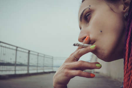 Young punk girl smoking cigarette. Hipster with pink dyed hair. Woman with piercing and unusual hairstyle. sadness, loneliness sadness on the face of a young informal model. subcultureの写真素材