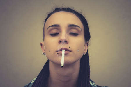 Beautiful young grunge rock girl smoking cigarette. Outdoor portrait of informal model. A woman with a piercing on her face, on the cheeks on her lips on her eyebrows. subcultureの写真素材