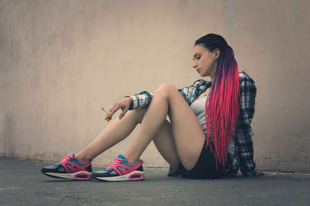 beautiful woman smokes a cigarette. informal model sitting on the pavement amid the old walls. Stress, hard life. problems in his personal life. loneliness, sadness, devastation. subcultureの写真素材
