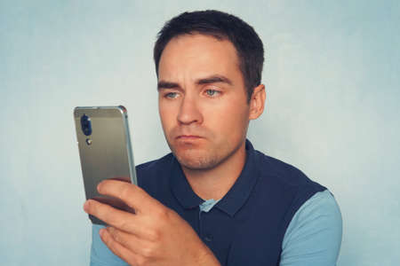 Disgusted man reading a text message. A sad expression on the face of a young man holding a modern smartphone on a blue background.の写真素材