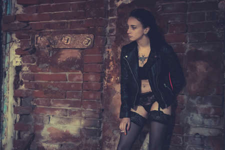 Sexy model stands old brick wall and poses in the image of a corrupt girl. Prostitute on the street smoking a cigarette with his knee bent. Girl addict in a leather jacketの写真素材
