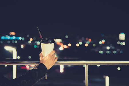 Closeup of female hand holding black craft coffee cup to take away, mock up of paper coffee cup to go outside, bokeh light. Night life style. paper Cup.の写真素材