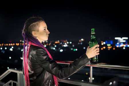 Portrait of lonely woman drinking alcohol from bottle at highway. A young drunk girl with a bottle of whiskey stands on the roof of the house and looks down at the night city.の写真素材