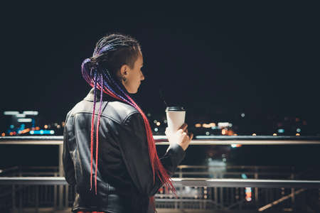 Beautiful young woman with coffee cup in the city. A thoughtful woman looks at the night lights of...