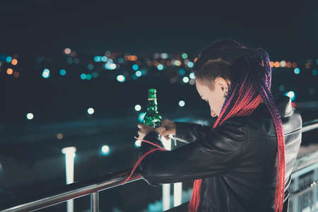 Girl drinking whiskey from a bottle in an attempt to forget. Drunk girl on the background of the city at night on a high floor looking down. atmosphere of sadness and sorrowの写真素材