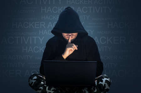 Man with hidden face working at a computer. Holding a finger to his mouth. Computer hacker stealing data from a laptop concept for network security, identity theft and computer crimeの写真素材