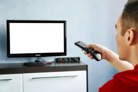 TV remote control in male hand in front of widescreen TV set with blank screen on blue wall background. Young guy switches channels on the remote from the TV.の写真素材