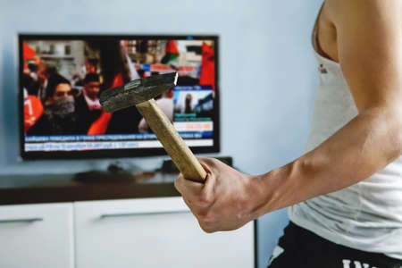man watches TV news report about the protests. angry guy smashes TV with hammer. false news caused anger among viewers. explosive nature. An angry viewer is dissatisfied with the report of the channelの写真素材