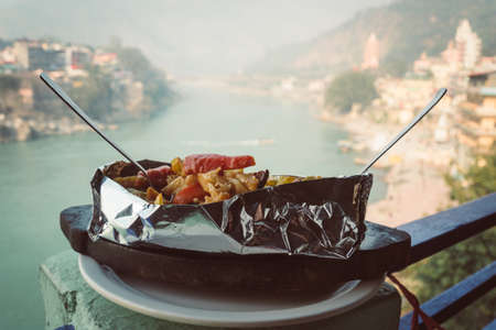 A dish of grilled and stewed vegetables in foil on the background of the river Ganges. Rishikesh...