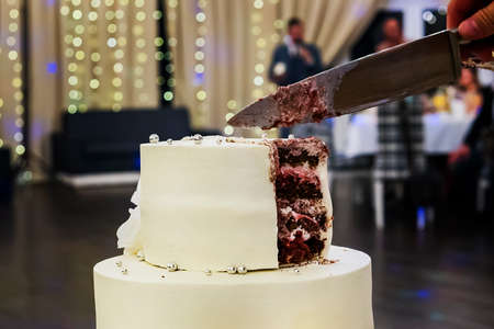 Cutting the first tier of a white mastic wedding cake in a banquet hall background. Wedding cake cutting sweet moment. Wedding traditions. cake etiquette. Ways of cutting a wedding cake.の写真素材