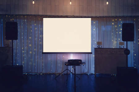 Illuminated banquette hall stage with a big white screen and acoustic systems. Equipment for video and audio projection at a festive event. Banquette stage with a board for projection in the centre.の写真素材