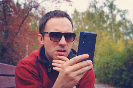 young man in wearing sunglasses holds a modern blue smartphone in his hand and look at the screen with a surprised look. Bewilderment on the face. The guy reads the message in the messengersの写真素材