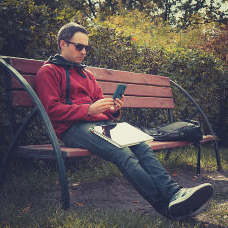Young hipster guy checking notification on mobile phone during distance work on laptop computer, sitting in city park in summer day.Handsome man chatting online on smartphone during webinar on netbookの写真素材