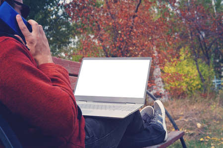 man working outdoors always connected with his smartphone and his loyal laptop. young guy, freelancer working outdoors in autumn Park. IT specialist at workの写真素材