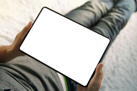 A lower half of a sitting man on the bed looking down to the empty screen of an up-to-date tablet. A...
