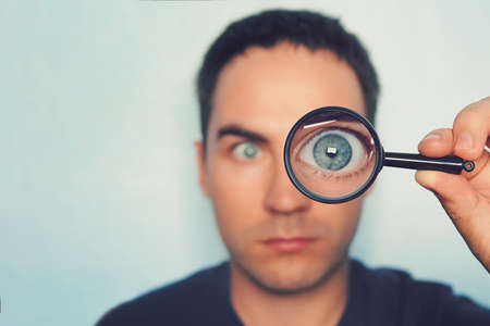 Potrait of young man looking through magnifying glass on white blurred background. View to male blue eye through the lens. Loupe with macro eye in persons hand on the foreground. Information search.の写真素材