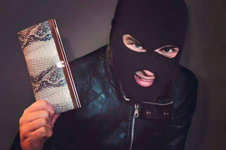 Masked criminal at the black background grins looking at the viewer and showing a purse in hand. Evil-doer is glad to have a stolen leather wallet. A satisfied robber after stealage. Thievish nature.の写真素材