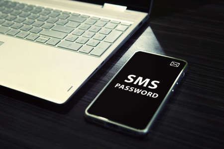 Table with open laptop and black-screened smartphone with white written words SMS password on its...