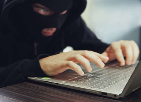 Close up of a ill-intentioned hacker in black balaclava entering the data on a laptop. Computer hacker commits cyber crime. A sitting man with hidden face types someting on the laptop. Internet crime.の写真素材