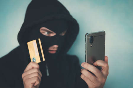 Close up of a man in robbery mask and hood holding the credit card and looking at the smartphone screen. Male criminal arranges a financial affair with mobile and credit card. Network fraud dangers.の写真素材
