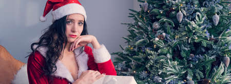 Sad girl sitting and thinking about real holiday. Serious and excited lady with a gift near the Christmas tree. copyspace for text and designの写真素材